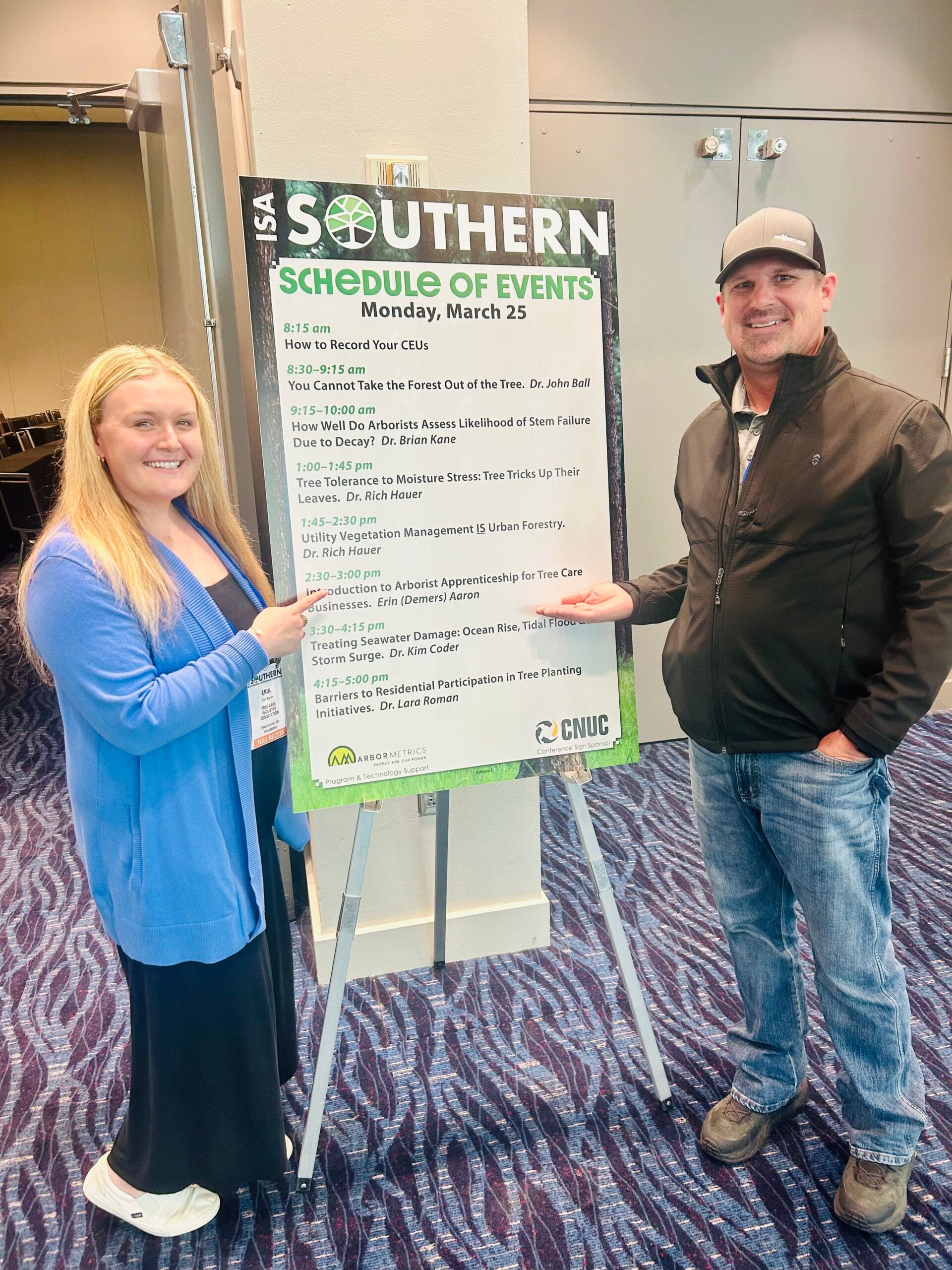 Woman and man standing beside a schedule of events board for the ISA Southern conference, featuring tree care topics and presentations.