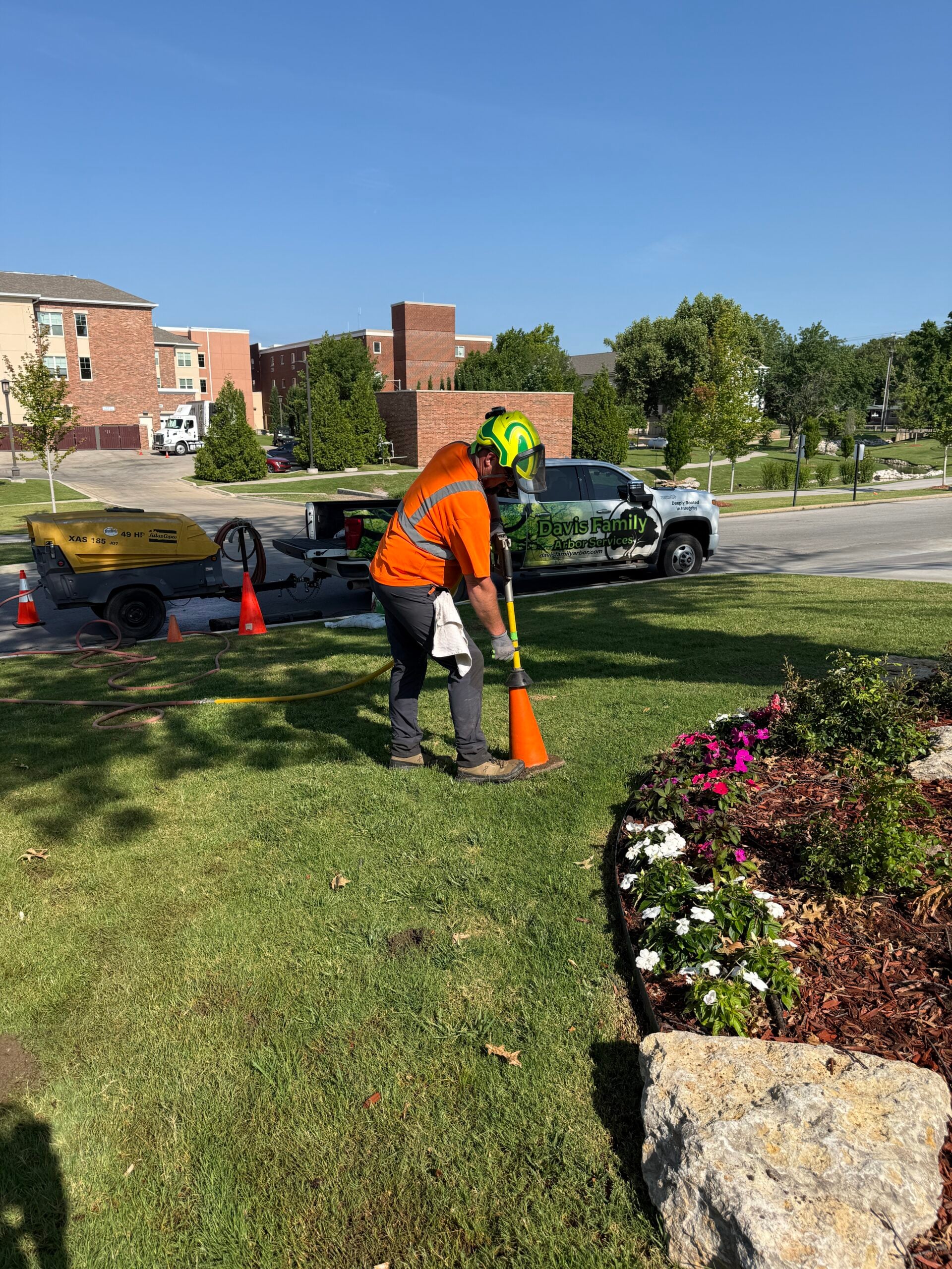 Arborist in safety gear using a tool near flower bed, with Davis Family Arbor truck in background, emphasizing tree care and preservation services.