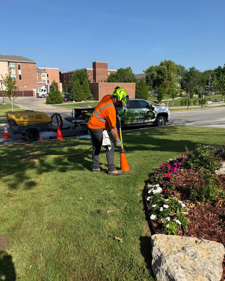 Arborist in safety gear using a tool near flower bed, with Davis Family Arbor truck in background, emphasizing tree care and preservation services.