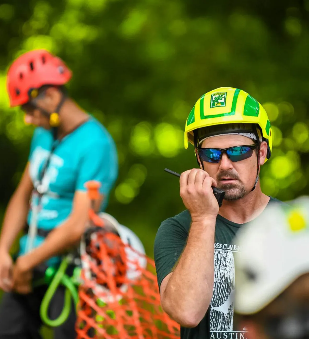 Tree climbing arborist in helmet and sunglasses speaks into a radio, with a fellow climber blurred behind him