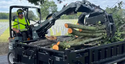Loader with a grapple loads cut tree logs into a dump trailer beside cemetery headstones
